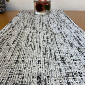 May include: A grey and white woven table runner with a subtle textured pattern. The runner is made from a soft, durable material and is perfect for adding a touch of elegance to any dining table.