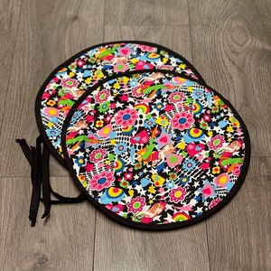 Op de afbeelding: Twee ronde zwarte en witte stoffen placemats met een kleurrijk psychedelisch patroon van bloemen, sterren en regenbogen. De placemats hebben zwarte banden.
