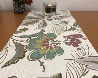 May include: A white table runner with a floral pattern in shades of brown, teal, and pink. The runner is decorated with a stylized floral design.