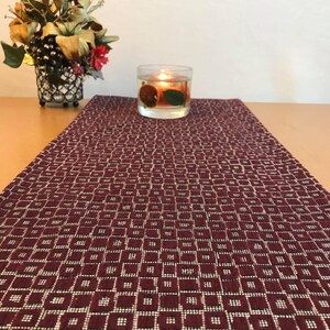 May include: A burgundy table runner with a white geometric pattern. The runner is on a wooden table with a clear glass candle holder with a lit candle and dried fruit inside.