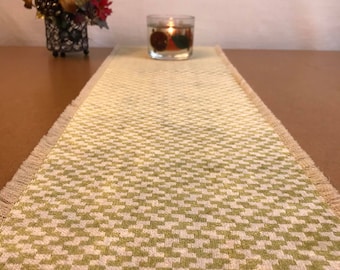 May include: A green and white checkered table runner with a fringed edge. The runner is on a wooden table with a candle in a glass jar in the center.