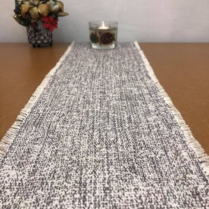 Peut inclure: Un chemin de table tissé blanc et gris avec des bords effilochés. Le chemin de table est sur une table brune avec un bougeoir en verre transparent au centre.