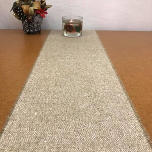 May include: A beige table runner with a herringbone pattern. The runner is on a brown table with a clear glass candle holder in the center.