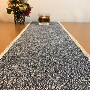 May include: A blue and white woven table runner with frayed edges. The runner is on a brown table with a candle in the background.