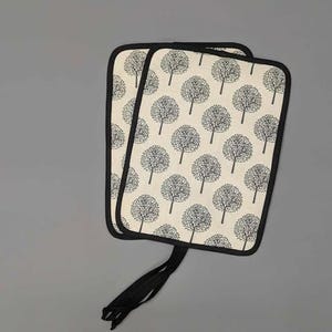 Puede incluir: Dos manoplas rectangulares con fondo crema y un patrón repetido de siluetas de árboles negros. Las manoplas tienen ribete negro y un lazo negro para colgar.