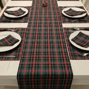 May include: A dining table set featuring a long rectangular table runner and six matching placemats, each with a dark green, black, and red plaid pattern. Six white plates are topped with folded plaid napkins and silver cutlery.