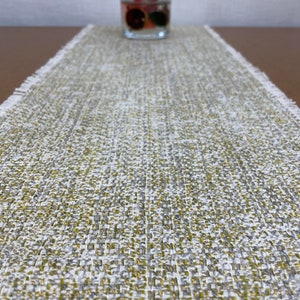 May include: A white and gold table runner with a textured woven design. The runner has a frayed edge and is ideal for adding a touch of elegance to any table setting.