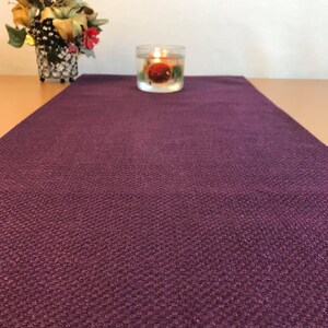 May include: A purple table runner with a textured surface. The runner is on a wooden table with a clear glass candle holder in the center.
