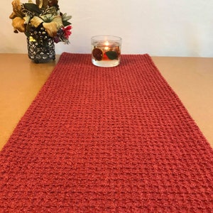 Peut inclure: Un chemin de table en tissu gaufré rouge avec un bougeoir en verre transparent contenant une bougie allumée et des fruits secs. Le bougeoir est placé sur le chemin de table.