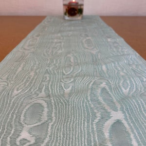 May include: A light green table runner with a wood grain pattern. The runner is made of a woven fabric and has a subtle sheen.