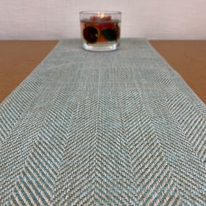Op de afbeelding: Een blauwe en witte visgraat tafelrunner met een teal en wit patroon. De runner ligt op een bruine tafel met een heldere glazen kaarsenhouder op de achtergrond.