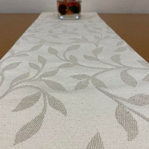 May include: A white table runner with a subtle leaf pattern. The runner is made of a woven fabric and has a slightly textured surface.