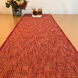 May include: A red and orange woven table runner with a subtle geometric pattern. The runner is fringed on the edges and is perfect for adding a touch of color and texture to your dining table.