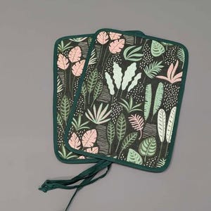 Può includere: Due presine verdi e nere con un motivo a foglie tropicali in rosa e verde. Le presine hanno un bordo verde e un laccio verde.