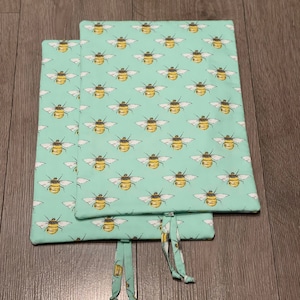 May include: Two rectangular fabric items with a light teal background and a repeating pattern of yellow and black bees with white wings. The items have matching fabric ties. The items are likely book covers or placemats.