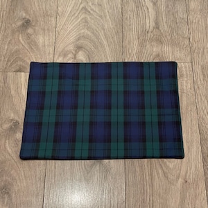 May include: A rectangular placemat featuring a traditional tartan design in navy blue, forest green, and black. The fabric has a woven texture, and the edges are neatly finished. This placemat is suitable for use at a dining table.