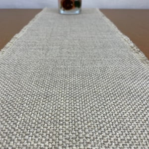 May include: A beige burlap table runner with frayed edges. The runner is textured and has a subtle sheen.