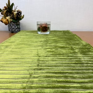 May include: A green velvet table runner with a striped pattern. The runner is on a wooden table with a clear glass candle holder in the center.