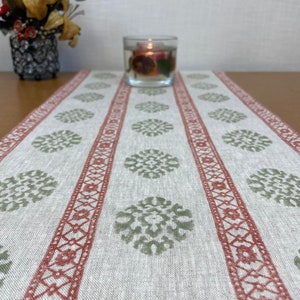 May include: A beige table runner with red and green geometric patterns. The runner has a subtle linen texture and features a repeating design of green circles with intricate details. The red stripes are bordered with a delicate, repeating pattern.