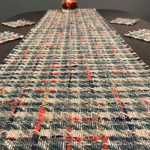 May include: A woven table runner with a blue, green and red plaid pattern. The runner is on a dark wood table with a candle in a glass jar in the centre.