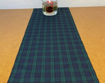May include: A navy blue and green plaid table runner. The runner is rectangular and features a classic tartan pattern. A glass candle holder with a lit candle and dried orange slice sits on the runner.