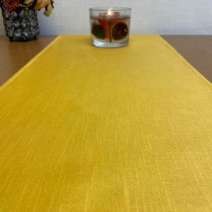 May include: A yellow table runner with a subtle textured pattern. The runner is on a brown table with a clear glass candle holder with a lit candle and fruit slices inside.