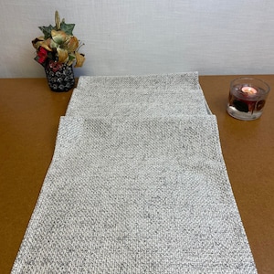 Può includere: Un runner da tavolo bianco e grigio testurizzato con un sottile motivo intrecciato. Il runner è disposto su un tavolo in legno marrone.