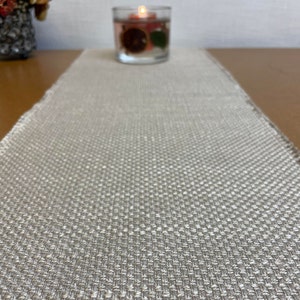 Op de afbeelding: Een beige linnen tafelkleed met een subtiel geweven patroon. De loper ligt op een houten tafel met een glazen kaarsenhouder in de achtergrond.