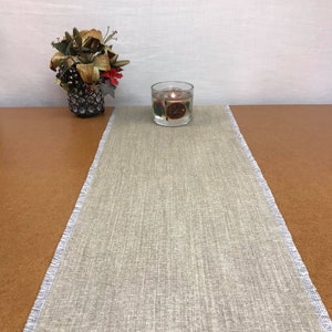 May include: A light gray linen table runner with a frayed edge. The runner is on a brown wooden table with a clear glass candle holder with a floating candle and dried orange slices.