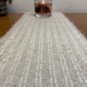 May include: A beige table runner with a white textured pattern. The runner has a frayed edge and is made of a soft, woven fabric.
