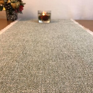 May include: A sage green table runner with a textured weave and fringed edges. A small glass candle holder with a lit candle sits on the runner. A small decorative floral arrangement is in the background.
