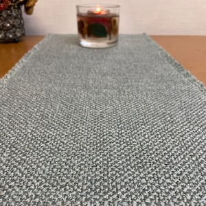 May include: A grey woven table runner with a subtle textured pattern. The runner is laid out on a wooden table with a clear glass candle holder in the centre.