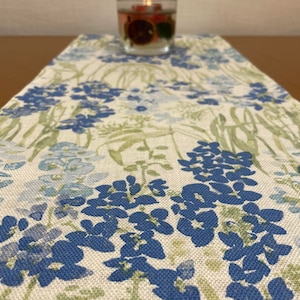 Op de afbeelding: Een bloemen tafelloper met een blauw en groen bloemmotief. De loper is gemaakt van een linnen-achtige stof en heeft een kaars in een glazen houder erop. De bloemen zijn helderblauw en de bladeren groen.