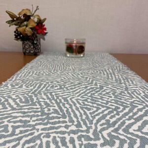 May include: A white table runner with a blue geometric pattern. The runner is made of a woven fabric and has a frayed edge. A clear glass candle holder with a lit candle is on the table runner.