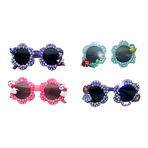 May include: Four pairs of flower-shaped sunglasses with rhinestones and character charms. The sunglasses are in purple, pink, blue, and teal.