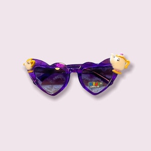 May include: Purple heart-shaped sunglasses with a yellow and white teapot and a yellow and white teacup on the sides. The sunglasses have a purple frame and lenses. The text "KIDS" is printed on the right lens.