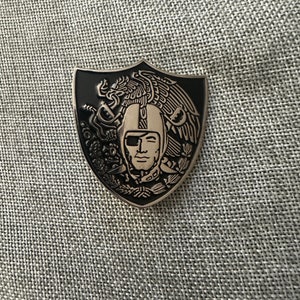 May include: A black and silver enamel pin featuring a stylized image of a football player's head with an eagle on the left and a serpent on the right. The pin is shaped like a shield.