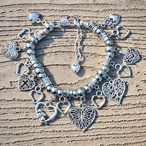 May include: A silver charm bracelet featuring a range of heart-shaped charms, including a butterfly, a cross, and a tree. The bracelet is made of silver beads and has a lobster clasp.