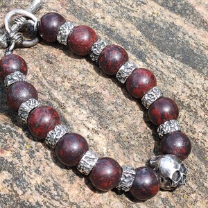 Könnte beinhalten: Ein Perlenarmband mit dunkelroten, runden Perlen und silberfarbenen Abstandshaltern. Das Armband hat einen silbernen Totenkopf-Anhänger und einen silbernen Verschluss. Der Schmuck wird auf einer strukturierten, braunen Steinoberfläche präsentiert.