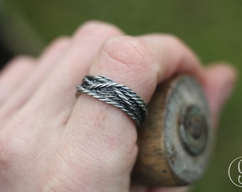 May include: A silver ring featuring a braided rope design. The ring is worn on a finger, with a blurred background. The ring's texture and design are the focal point, showcasing a unique, handcrafted jewellery piece.