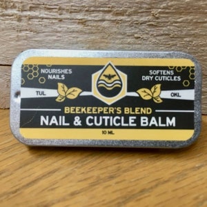 May include: A silver tin with a yellow and black label that reads "BEEKEEPER'S BLEND NAIL & CUTICLE BALM 10 ML". The label also includes the text "Nourishes Nails", "Softens Dry Cuticles", "TUL", and "OKL".