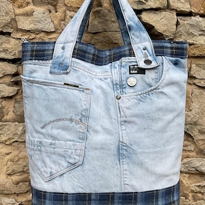 May include: A light blue denim tote bag with a plaid fabric trim. The bag has a large pocket on the front and a smaller pocket on the back. The bag has a G-Star RAW logo on the front.