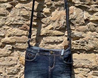 May include: A dark blue denim crossbody bag, designed to resemble jeans, with pockets and white stitching. The bag has a black strap and a decorative button. It hangs against a stone wall.