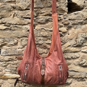 Puede incluir: Un bolso hobo de color rosa empolvado con una correa vertical central de cuero marrón oscuro y cierre de hebilla metálica. El bolso incluye dos bolsillos frontales con cremallera y herrajes plateados, una correa ancha y costuras visibles.
