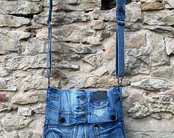 May include: A blue denim crossbody bag constructed from repurposed jeans, featuring a front flap with visible metal buttons, a small pocket, and an adjustable denim shoulder strap. The design incorporates classic jean pocket details and seams.