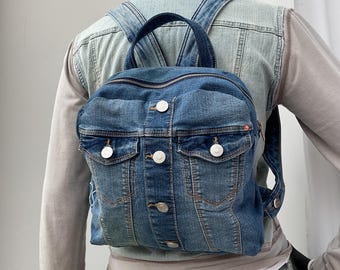May include: A blue denim backpack with a front pocket design, featuring silver-colored buttons and a top handle. The backpack has adjustable straps and is made from a faded denim material.