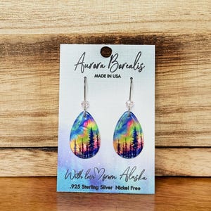 May include: Teardrop-shaped earrings with a colorful aurora borealis design and a forest silhouette. The earrings are suspended from silver hooks with a small clear bead. The card reads "Aurora Borealis, Made in USA" and "With love from Alaska."