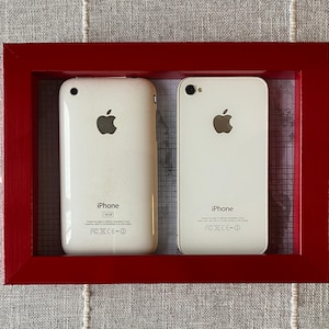 Puede incluir: Dos teléfonos iPhone blancos se muestran en un marco rojo. Los teléfonos están mirando a la cámara y muestran la parte posterior de los teléfonos. Los teléfonos tienen el logotipo de Apple y el texto "iPhone" en la parte posterior.
