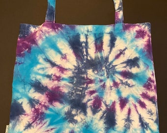 May include: A blue, purple and white tie-dye tote bag with a circular pattern. The bag has two long straps.