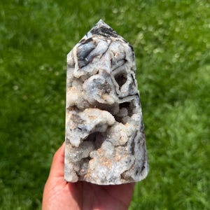 May include: A natural crystal tower with a pointed top. The crystal features a mix of white, gray, and black colors, with a textured, porous surface. The crystal is held in a hand against a backdrop of green grass.